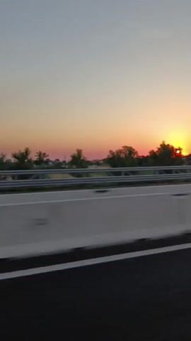 The video shows a view from a moving vehicle, likely a car, looking out at a sunset. The foreground features a highway barrier and a lane marking. In the background, a line of trees is silhouetted against the orange and yellow hues of the setting sun. The sky transitions from a pale blue at the top to warm colors near the horizon. There are no people visible in the video. The overall impression is one of travel and a scenic sunset.
