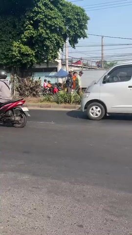 The video shows a street scene during the day. In the foreground, a motorcycle with a rider is moving away from the camera on the left side of the asphalt road. On the right side of the road, a silver minivan is stopped, facing right. In the background, several people are gathered near a sidewalk. Some are standing, and others are on motorcycles. There is a blue tent set up behind them, and a large tree with green foliage is to the left of the gathered people. Power lines are visible in the sky. The overall environment appears to be an urban or semi-urban street.