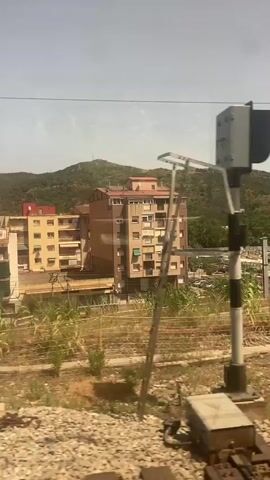 The video shows a view from a moving train. In the foreground, there are train tracks and gravel. Beyond the tracks, there is a patch of dry, brown ground with some sparse green vegetation. Further back, there are tall green plants and then a concrete barrier with some construction materials, including pipes and yellow electrical boxes. To the right, there is a tall pole with graffiti on it and more train tracks. In the middle ground, there are several multi-story apartment buildings. The buildings are a mix of beige and brown, with visible windows and balconies. Behind the buildings, there is a large parking lot filled with cars. Further in the background, there are rolling green hills covered in trees, with some structures visible on the hilltops, possibly antennas or towers. The sky is clear and blue with some hazy white clouds. The sun appears to be shining brightly.