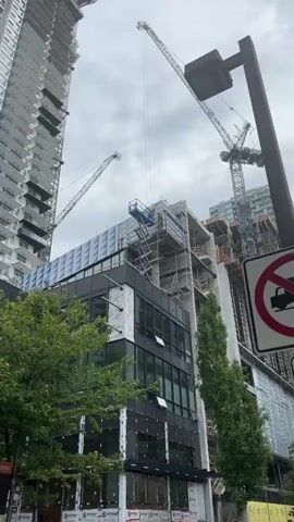 This video captures a daytime scene at a construction site in Coquitlam, Canada, under an overcast sky. Two construction workers are actively involved in installing facade panels on the upper levels of a building.

One worker is operating a blue Genie GS-3232 scissor lift, positioned high against the building's exterior. The other worker is on scaffolding directly next to the building, assisting with the installation. Both individuals are wearing essential safety gear, including hard hats and high-visibility vests.

The building features a modern design, with finished dark-framed windows on its lower floors. The section where the workers are concentrated is in an earlier stage of completion, with structural concrete elements visible alongside recently installed white panels. These panels display the brands "SOPREMA" and "SOPRASEAL."

In the background, two prominent construction cranes tower over the site, indicating a large-scale development project. Green tree foliage is visible at the bottom of the frame, grounding the industrial scene. The workers appear focused on their task, meticulously securing the building materials.