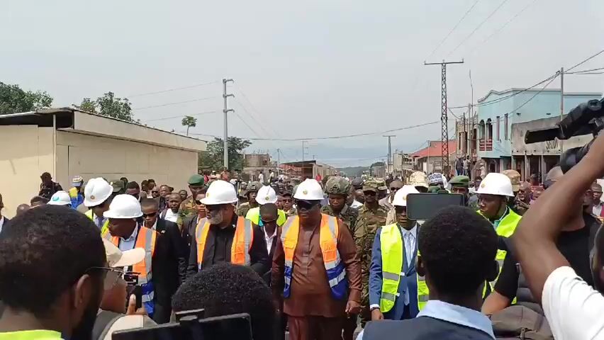 A crowd of people wearing white hard hats and bright orange safety vests walks down a street in an urban area. Some people are wearing military uniforms. The sky is overcast, and buildings line the street. There are power lines overhead. Some people in the crowd are wearing sunglasses. One person is holding a camera. The camera moves with the crowd.
