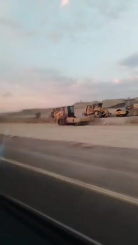 The video shows a construction site from a moving vehicle. Several bulldozers are visible on a dirt path, some of them moving earth. The terrain appears to be hilly and dry, with some vegetation scattered throughout. The sky is partly cloudy with a mix of blue and pink hues, suggesting it might be sunrise or sunset. The road in the foreground is paved, with a yellow line visible. The footage is somewhat blurry due to the movement of the vehicle.