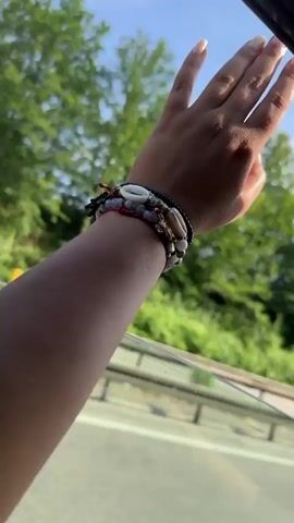 The video shows a person's arm extended outside of a car window as the car is moving. The arm is adorned with multiple bracelets of varying designs and materials. A bright blue sky is visible overhead, with some white clouds. Lush green trees line the roadside, forming a blurred backdrop as the vehicle speeds along. The interior of the car is partially visible, showing the edge of the car door frame and a portion of the window glass. Other vehicles can be seen on the road, including a white van that briefly passes by. The person's hand is outstretched, fingers splayed, as if feeling the wind or reaching towards the landscape.