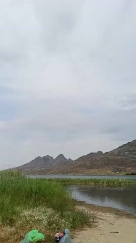 A video shows a scenic view of a lake with mountains in the background. There are reeds growing along the shore, and the water is calm. The sky is overcast, with a few clouds scattered across it. In the foreground, there is a sandy beach with some camping gear, including two green sleeping bags and a blue bag. There are also a few small buildings visible on the far shore of the lake, nestled against the foothills of the mountains. In the water, a person can be seen swimming. The video captures a peaceful, natural environment, perfect for outdoor recreation.