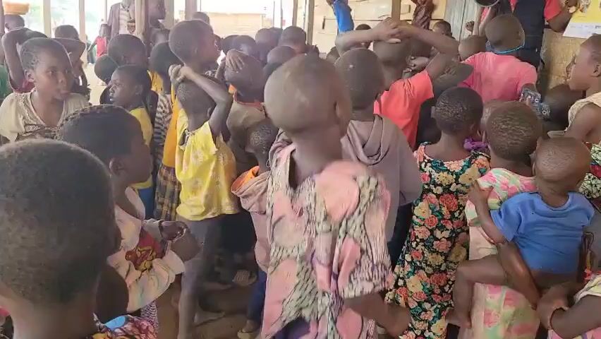 The video shows a crowd of children gathered in an open-air, possibly rural, setting. The children appear to be of various ages and are dressed in casual clothing. Some are looking around, while others are interacting with each other. The environment seems to be a gathering space, possibly a school or community center, with wooden structures and a dirt floor. The overall atmosphere appears to be one of a group activity or assembly.