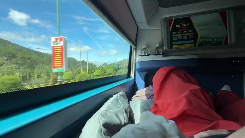 The video shows a view from inside a moving vehicle, possibly a bus, looking out a window. The window has a blue frame and a shaded screen at the top. Outside the window, there is a landscape with green trees and rolling hills under a bright blue sky with scattered white clouds. A road with some traffic can be seen. Inside the vehicle, there are beds or sleeping compartments, with blankets and pillows visible. One person is covered by a red blanket. A digital screen is mounted on the wall of the vehicle's interior, displaying text and images. Two small bottles are also visible on a shelf above the beds. The overall impression is that of a passenger traveling in a sleeper bus or similar vehicle.