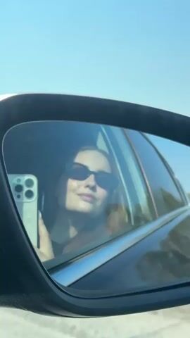 The video shows the reflection in a car's side mirror of a woman wearing sunglasses, holding a phone. The sky is clear and blue. The car appears to be moving on a road, with glimpses of trees passing by in the background. The woman is looking towards the camera, likely taking a selfie. The phone appears to be an iPhone.