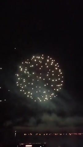 The video shows a fireworks display at night. The fireworks are various colors and shapes, and they are exploding in the sky. There are also some people in the foreground, holding up their phones to record the fireworks. The fireworks are illuminating the clouds and the water below. The scene is festive and celebratory.