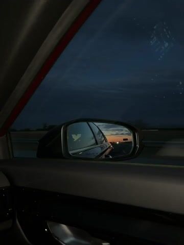 The video shows a view from inside a car, looking out the side window and into the side mirror. The sky is dark blue with clouds, suggesting dusk or night. In the side mirror, a sunset is visible with orange and yellow hues, reflecting off the car's window. The car appears to be moving on a highway, indicated by the blurred landscape in the background and the perspective of the side mirror. A faint reflection of a bird or wing-shaped logo is visible in the side mirror. The interior of the car is dark and out of focus, with a red trim visible on the window frame.