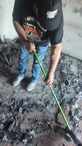 The video captures an overhead view of an individual engaged in cleanup within a fire-damaged interior space in Lirquen, Chile. The person is wearing a black t-shirt, blue jeans, and light-colored shoes, and is actively sweeping or pushing burnt debris and ash across the floor using a broom with a green handle and purple bristles.

The scene depicts extensive fire damage. The floor is covered in a thick layer of dark ash, charred wood fragments, and various pieces of broken, burnt material. Patches of the underlying floor are visible, showing what appears to be a concrete or tiled surface. The surrounding walls exhibit peeling paint and significant soot staining, further indicating the severity of the fire. Among the debris, a damaged, twisted metal object can be seen, possibly remnants of furniture or an appliance.

The activity clearly shows the early stages of recovery and restoration after a fire. The individual's movements are purposeful, focusing on clearing the destructive aftermath.

Visible text on the person's black t-shirt reads: "I NEVER DREAMED I'D GROW UP TO BE A SUPER SEXY SHEET SHOW ADVISOR". Additionally, part of the word "ENGINE" is visible on what appears to be a cap or another part of their attire. The time of day is not discernible from the indoor setting, which appears to be lit by ambient or artificial light.