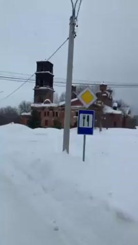 The video shows a partly ruined brick building in a snow-covered landscape. The building features a tall tower with visible structural damage and missing windows. The sky is overcast, indicating a winter or cold weather setting. A road sign and power lines are visible in the foreground. The camera appears to be moving, suggesting the scene is viewed from a vehicle passing by.