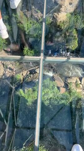 The video is shot from a top-down perspective looking through a glass floor. Below the glass floor is a natural landscape with trees, rocks, and a river or stream with a waterfall. Several people are visible walking on bridges and pathways below, as well as on the glass walkway. The perspective shifts slightly as the camera moves, revealing different sections of the scene. The feet of at least two individuals wearing white sneakers are visible in the foreground, standing on the glass floor.