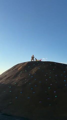 The video shows a man standing on top of a climbing hill at a playground, holding a young child above his head. The man is wearing a t-shirt and shorts. The child is wearing pants and a jacket. The man lowers the child back down and stands over them protectively. The sky behind them is clear and blue. The climbing hill has colorful climbing grips attached to its surface. Some other playground equipment is visible in the background.