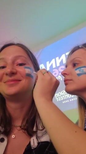 Women apply Israeli flag face paint near Eurovision Vienna signage