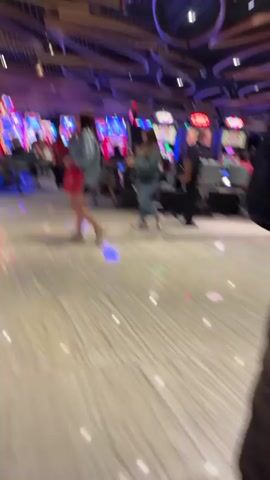 The video shows a wide-angle view of a casino floor. Several people are walking around, some are seated at slot machines. The floor is made of polished marble with a red and white pattern. The ceiling is high and features a modern design of interconnected wooden elements and purple lights.  A man in a white shirt and black vest walks towards the right of the frame.  The video is shot from a moving perspective, suggesting the person filming is walking.