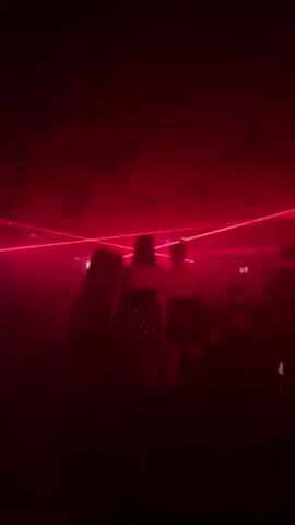A low-angle, hand-held video shows a crowd of people dancing at a club. The room is dark, illuminated by vibrant red laser lights that sweep across the ceiling. The people are silhouetted against the light, and one woman is wearing a dress with white polka dots. The camera shakes slightly as the person filming moves around.