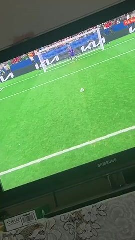 The video shows a television screen displaying a soccer match. A player in a red jersey with the name "M. SALAH" and the number "10" on the back is visible. The player is in the process of kicking a soccer ball towards a goal defended by a goalkeeper in a purple jersey. The scene appears to be a penalty kick or a similar decisive moment in the game, as indicated by the close-up on the player and the intense action. The background shows spectators in the stands and advertising boards around the stadium. The overall environment is that of a professional soccer game being broadcast on television.