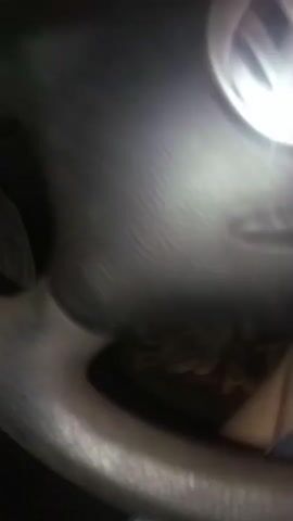 This is a close-up, low-angle shot of a person's hands operating a car's steering wheel and gear shift. The steering wheel is made of dark, textured material. A hand is visible on the left side of the steering wheel, operating the turn signal. Another hand is visible on the right side, operating the gear shift. The car's dashboard and speedometer are partially visible in the background. The lighting is dim, suggesting it is nighttime or a dimly lit interior. The video captures the actions of driving or preparing to drive.