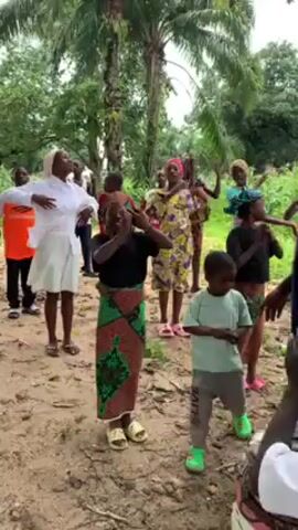 A group of children and adults are dancing outdoors in a wooded area.  They are moving their arms up and down, some are smiling.  There are palm trees visible and the ground is sandy. A man wearing a navy blue shirt walks through the group.  Several people are wearing traditional African clothing.