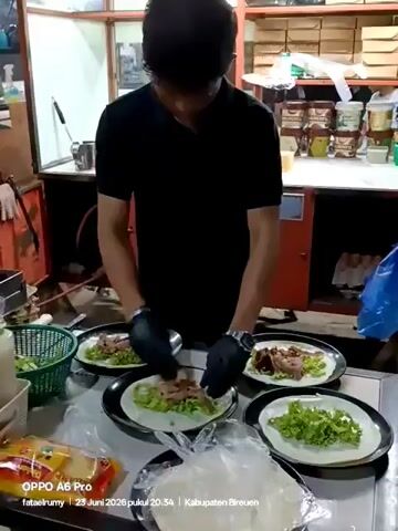 A person wearing black gloves and a black t-shirt is preparing food at a counter. There are four plates in front of the person, each containing a tortilla base with lettuce and sliced meat. The person is adding more meat to the tortillas. To the left of the plates is a basket of lettuce and various bottles and containers. To the right, there are stacks of tortillas wrapped in plastic. The background appears to be a kitchen or food stall with shelves filled with jars and containers. A timestamp overlay indicates "23 Juni 2026 pukul 20.34" and "Kabupaten Bireuen".