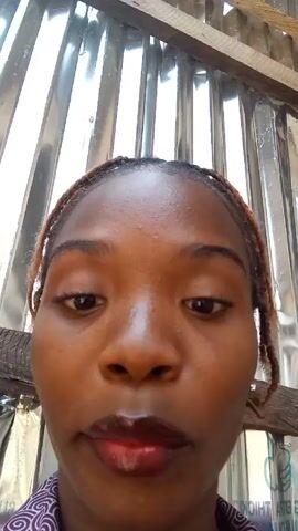 A medium close-up shot of a person facing forward is positioned below their forehead to the middle of their chest. The person has brown skin, brown eyes, thin lips painted with red lip gloss, and thin brown braids pulled back off their forehead. They are wearing a purple patterned shirt. The background is a corrugated metal wall or roof.