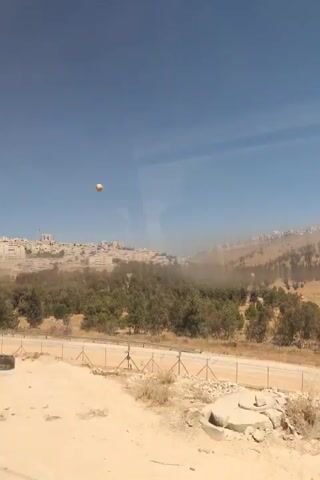 The video shows a dusty, rocky landscape with a cityscape visible in the distance on a hillside under a clear blue sky. In the foreground, there is a tall metal utility pole with electrical equipment attached. A tire is lying on the ground near the pole. A bright orange object, possibly a ball, is visible in the air in the upper right quadrant of the frame. The camera appears to be moving, possibly from a vehicle, as indicated by the slight blur and the changing perspective. There are no people or direct human interactions visible in the frame.