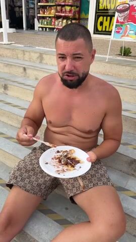 A man with short dark hair and a beard sits on concrete steps outdoors. He is shirtless and wearing patterned shorts. He holds a white paper plate with a fork in his left hand and a fork in his right hand. The plate contains a dessert with chocolate sauce and some white powdery substance. The man is looking at the camera. Behind him, there are steps, shops, cars, trees, and a cloudy sky.