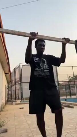 A young man is attempting pull-ups on a wooden bar outdoors. He is wearing a black t-shirt and black shorts, and he is barefoot, wearing slide sandals. He has a beard. He struggles to pull himself up.  The bar is supported by what appear to be makeshift supports. The background features a tiled area, a building, a tennis court with a net, and greenery. The sky is visible.