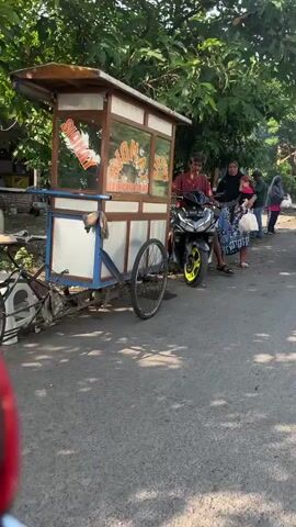A motorcycle is parked on the side of a road. A person is sitting on the motorcycle. Another person, wearing a headscarf, is standing next to a food cart. A woman is walking with a child. There are trees and foliage in the background. The ground is paved. The scene appears to be outdoors during the daytime.