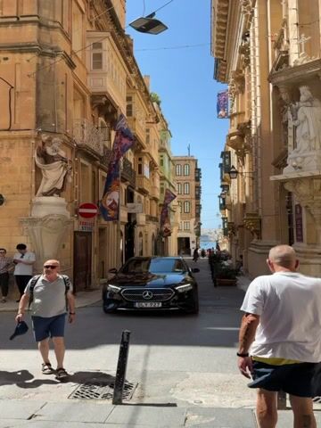 The media file captures a sunny day in Valletta, Malta, showcasing a narrow, historic street lined with traditional Maltese architecture. The buildings feature the distinctive enclosed wooden balconies, some painted in vibrant green, others in earthy yellow and beige tones, stacked one above the other. Ornate carvings and statues adorn the facades, hinting at the area's rich history.

In the foreground, two men are walking away from the camera. The man on the left wears a grey t-shirt, blue shorts, sunglasses, and a backpack, while the man on the right sports a white t-shirt and dark shorts. They appear to be tourists enjoying the city.

The central focus of the street is a sleek, black Mercedes-Benz car. Its license plate reads "DLY 927". The car is stationary, likely waiting for traffic or navigating the tight street. Several people can be seen in the distance, walking towards a sunlit harbor or waterfront visible at the end of the street, suggesting a lively atmosphere.

Large, colorful banners with elaborate designs hang from the buildings, adding a festive or decorative element to the scene. One banner prominently displays a crest with red and gold elements. A red and white circular traffic sign is also visible on the left, indicating a prohibition. Below it, a sign for "FRANKNER STORE" is partially obscured.

The clear blue sky and bright sunlight indicate it is midday or early afternoon, and the weather is warm. The overall impression is that of a picturesque and well-preserved European city, popular with tourists, where modern life, represented by the car, coexists with ancient surroundings. The scene evokes a sense of exploration and the enjoyment of cultural heritage under pleasant weather conditions.