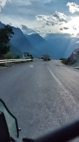 A low-angle video captures a view from a vehicle driving on a paved mountain road. The sun shines through dramatic clouds, casting rays over a vast mountain range in the distance. Two white vans and one white car are parked or driving on the road. A sign is visible on the left side of the road, featuring text and an arrow. The road curves to the right as the vehicle continues its journey.