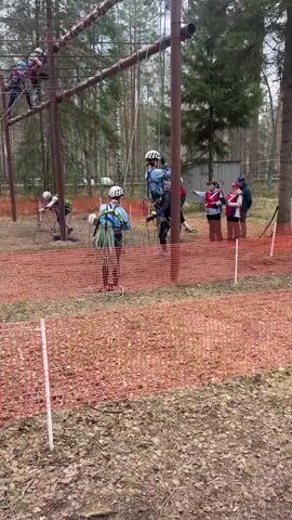 The video shows a group of people in a wooded area, engaged in what appears to be a ropes course activity. Several individuals, wearing helmets and harnesses, stand near the ground, while one person is navigating a structure constructed from poles and ropes overhead. There is a barrier of orange mesh fencing around the perimeter of the activity area, possibly defining the course boundaries or providing a safety perimeter. Several of the people on the ground are wearing red and white vests, and some appear to be pointing or instructing. The ground is covered in dried leaves and reddish-brown dirt or gravel. The surrounding environment consists of tall trees.