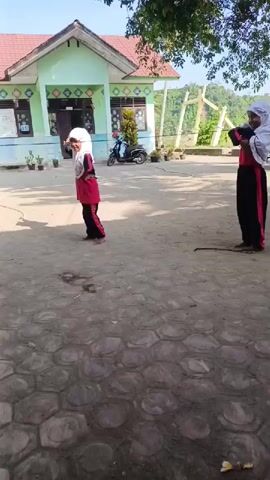 Two young girls are playing an outdoor rope game on a sunny day, likely in a schoolyard or community area in Simpangjaya, Indonesia. One girl, wearing a red and black outfit with a white hijab, is actively jumping over a rope that is being swung by the other girl, who is wearing a white hijab, red top, and black pants. Both girls appear happy and are smiling as they play. In the background, a light green building with a red roof is visible, along with a parked scooter, potted plants, and trees. The ground in the foreground is paved with hexagonal stones.