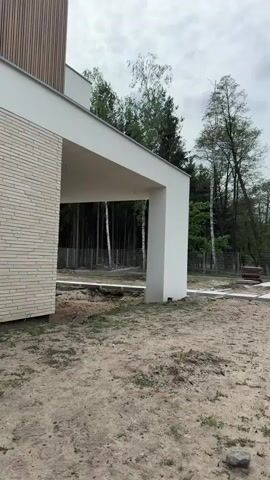 The video shows the exterior of a modern house under construction. The house has a white cantilevered roof supported by a white pillar, and a brick facade on one section. The surrounding ground is mostly bare dirt with some patches of grass. In the background, there is a fence bordering a forest with a mix of deciduous and evergreen trees. The sky is overcast. The camera pans slightly from left to right.