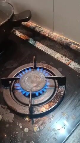 A close-up video shows a gas stovetop with a lit burner. The burner emits a blue flame. A wooden spatula is being held above the flame. The stovetop is black with some food residue visible. The countertop behind the stovetop is a dark color with light-colored flecks, and the backsplash is made of light-colored tiles.