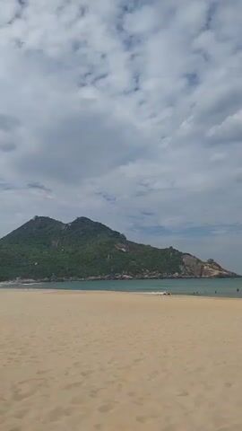 A wide sandy beach stretches into the foreground of the frame, with the ocean and a large, tree-covered mountain in the background. The sky above is filled with white and grey clouds. A few people are scattered along the shoreline, some wading in the water and others sitting on the sand. The waves gently roll onto the beach. The overall scene is peaceful and natural, bathed in soft, diffused light.