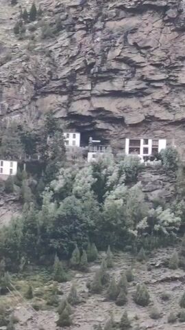 This video pans across a steep, rocky mountainside. Several structures, appearing to be buildings, are built into the cliff face or on ledges. Some of these structures are white with dark window frames, while others have a more rustic appearance with wooden elements. Lush green trees and vegetation grow at the base of the mountain and on some of the ledges. The terrain is rugged and uneven, with patches of sparse vegetation and scattered rocks. In the distance, a wider view of the valley reveals more structures, including what appear to be homes or a village, situated on a gentler slope. Terraced fields or gardens are visible on the lower slopes. The overall impression is of a remote, mountainous region with human habitation integrated into the challenging landscape.
