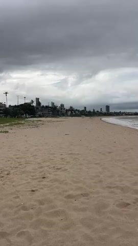 The image depicts a beach scene in João Pessoa, Brazil, under a cloudy sky. The foreground is dominated by a sandy beach with visible footprints, suggesting recent activity. The middle ground features the ocean with gentle waves lapping at the shore. In the distance, a cityscape with several tall buildings lines the coast, interspersed with palm trees. The sky is filled with heavy, dark clouds, indicating overcast weather, possibly before or during a rain shower. The overall mood is somewhat somber due to the weather, yet the presence of the beach and city suggests a populated area, though no people are clearly visible in the frame. The time of day appears to be daytime, but the cloudy conditions obscure direct sunlight. There is no visible text in the image.