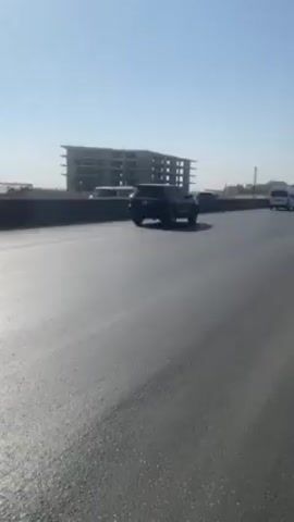 The video shows a straight highway on a sunny day.  A partially-constructed building is visible in the distance. A white van drives in the distance. Two trucks are parked on the right side of the road, and another large orange truck drives past.