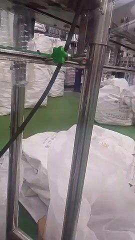 The video captures an overhead view of a conveyor belt system within a warehouse or factory setting. Large, white, opaque bags are stacked in rows on the floor, with some partially obscured by the industrial equipment in the foreground. The equipment consists of silver-colored metal poles and a green knob attached to a dark cable, which appears to be part of the conveyor mechanism. The bags are uniformly sized and appear to be filled with a granular or powdery substance, as suggested by their shape and the slight bulging. The overall environment suggests an industrial or manufacturing facility focused on bulk material handling and packaging.