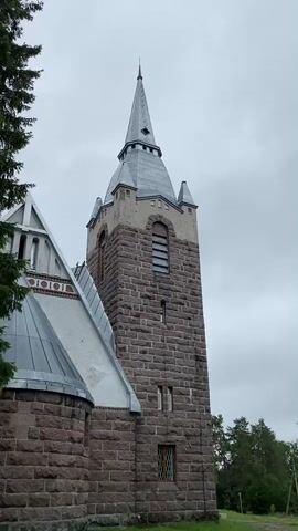 The video shows a tall, stone church tower with a spire against a cloudy sky. The tower is constructed of large, rough-hewn stones in shades of brown and grey. It has small windows and decorative architectural elements. To the left of the tower, a portion of the church's roof and side wall is visible, also made of stone with a metal roof. A large evergreen tree stands to the far left of the frame, partially obscuring the church. The ground in front of the church is covered in green grass. The camera slowly pans slightly to the left over a period of four seconds.