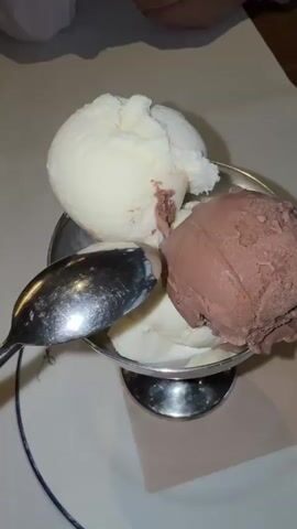 A close-up video shows a dessert bowl containing two scoops of ice cream: one white (vanilla) and one brown (chocolate). A silver spoon rests in the bowl, partially in the white ice cream. The bowl is placed on a white plate with a blue rim, which also features a light-colored napkin underneath. Faint, illegible text is visible on the plate. The lighting in the scene is dim, suggesting an indoor setting like a restaurant or cafe.
