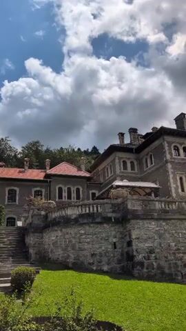 A video pans across a large stone building with a red-tiled roof, set against a backdrop of trees and a cloudy sky. The building appears to be a mansion or a historical structure, with multiple stories and ornate architectural details, including arched windows and balconies. A stone wall surrounds the lower portion of the building, and a well-maintained lawn is visible in the foreground. A tall, slender tree stands to the right of the frame, partially obscuring the building. The overall scene suggests a serene and picturesque setting, possibly a tourist attraction or a private estate.