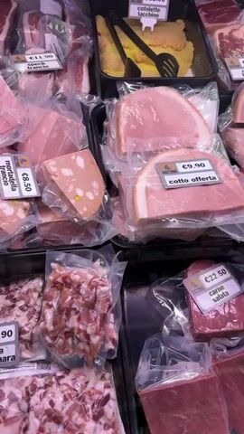 The video shows a display of various types of cured meats and sausages in a shop. The camera pans across rows of packaged products, some of which have price tags. Visible items include mortadella, pancetta, cured sausages, and what appear to be various types of salami. Some of the sausages are hanging, while others are packaged in trays. The lighting suggests an indoor setting, possibly a deli counter or a market stall. The camera movement is smooth, indicating a deliberate recording of the merchandise. No people are visible in the frame.