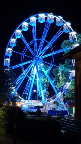 A Ferris wheel is lit up at night with changing colored lights. The Ferris wheel is large and has many gondolas. The lights on the Ferris wheel change from blue to pink, red, and purple. The Ferris wheel is surrounded by trees and a building. There are some people visible near the base of the Ferris wheel.