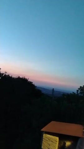 A time-lapse video of a sunset over a landscape of trees and hills. The sky transitions from a deep blue to lighter shades of blue and pink as the sun sets. The silhouette of trees is visible in the foreground, and a distant tower can be seen in the background.