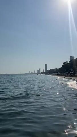 The video opens with a shot of the ocean on a clear, sunny day. The sun is shining brightly, and its rays are reflecting off the water, creating a sparkling effect. In the distance, a coastline is visible, lined with tall buildings and lush green trees. A sandy beach stretches along the shore, with a few people and beach umbrellas scattered about. Gentle waves are lapping at the beach, and the water appears calm and inviting. The camera pans slightly to the right, offering a wider view of the scene. The overall impression is one of a serene and beautiful coastal landscape.