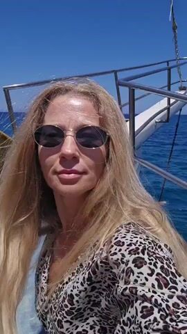 A blonde woman wearing sunglasses and a leopard-print top smiles at the camera. She is on a boat with the ocean and sky visible in the background.