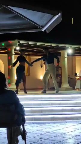 A man and a woman are dancing on a stage at night. The woman is wearing a blue dress and the man is wearing a blue shirt and white pants. The stage is lit with white lights and there are colorful lights on the columns. There are tables and chairs in the background. A person is sitting in a chair in the foreground, watching the dancers.