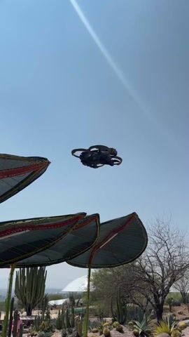 The video shows a drone flying in the sky on a sunny day. The drone is black and has four rotors. It flies over a park-like area with trees, plants, and buildings. In the distance, there is a cell tower. The video ends with a view of the park.